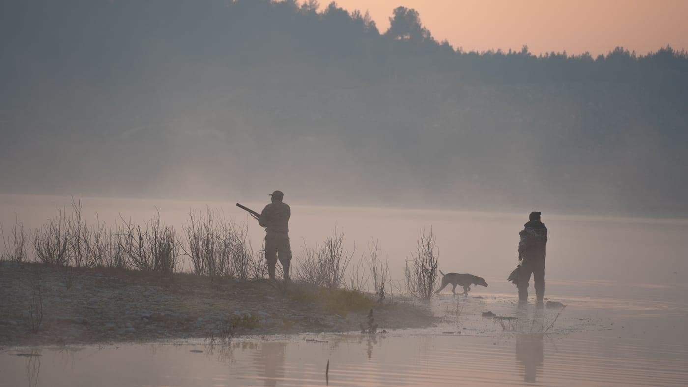 Hunters and a dog by a foggy lakeshore at sunrise