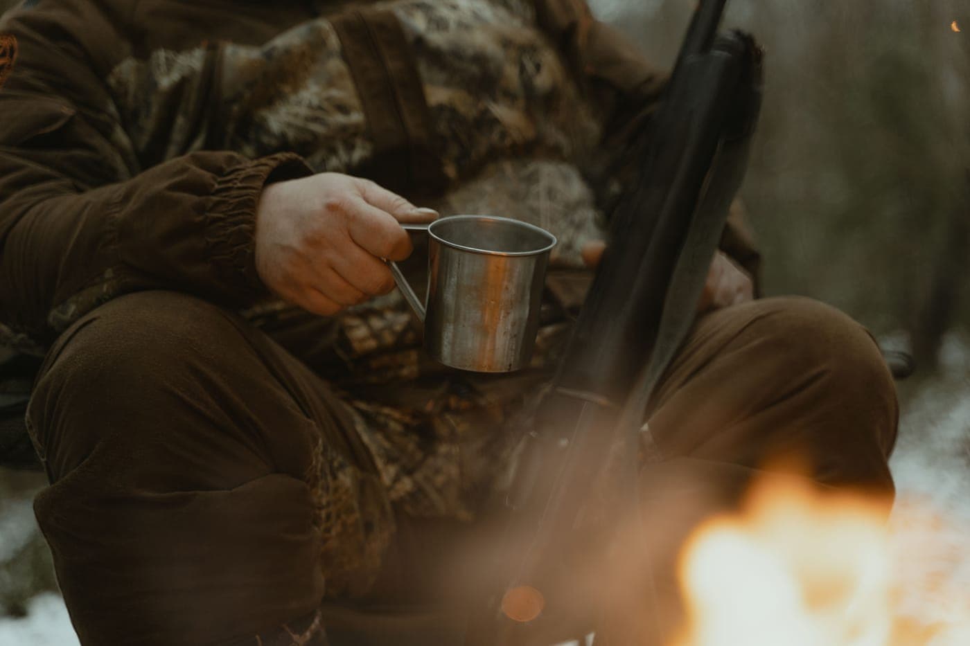 A person in camouflage sitting by a campfire with a tin cup